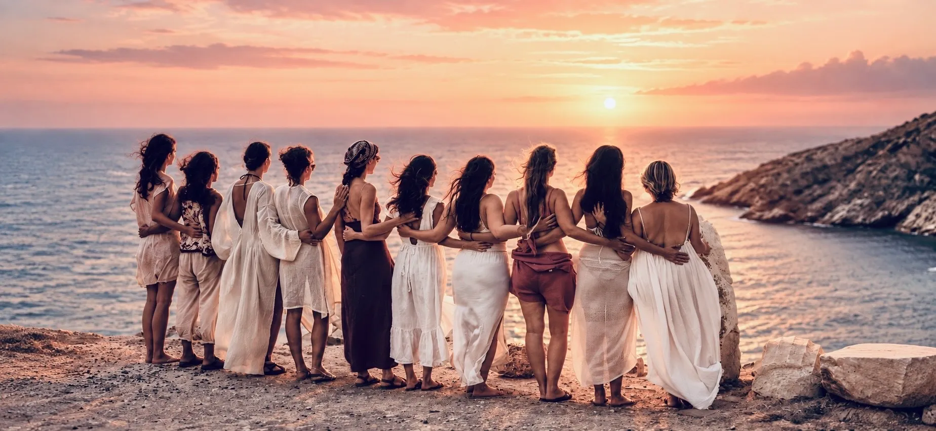 A circle of women with their arms around each other, looking out over the sea at sunset